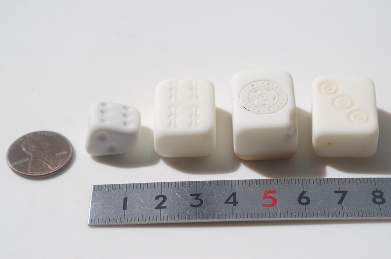 May include: A collection of small, white game pieces, including a die and several rectangular tiles, are arranged on a white surface. A ruler shows measurements in inches. A penny is also included for size comparison.