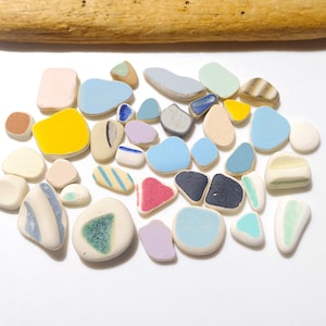 Peut inclure: Une collection de pierres en céramique lisses et colorées, de formes et de tailles variées, dont le bleu, le jaune, le rose et le vert. Un morceau de bois flotté est en haut. Les pierres sont disposées sur une surface blanche.