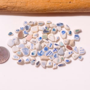 Op de afbeelding: Een verzameling kleine, gladde, witte keramische scherven met blauwe, bruine en groene patronen, verspreid naast een koperkleurige Amerikaanse penny voor schaal. De stukjes zijn onregelmatig van vorm en hebben diverse beschilderde ontwerpen en verweerde randen.