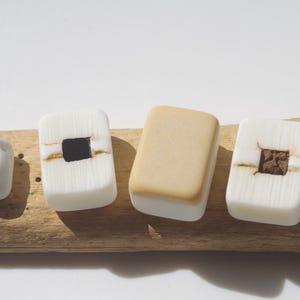 May include: Four decorative, handcrafted soap bars in various shapes and colors. One is a light gray, irregular shape. Two are white with a square black or brown center. The fourth is a tan rectangle. All are on a piece of driftwood.