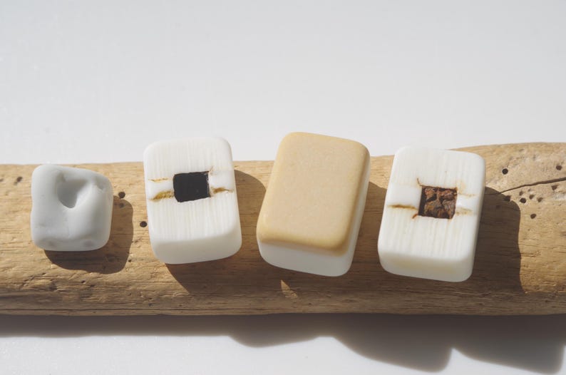 May include: Four decorative, handcrafted soap bars in various shapes and colors. One is a light gray, irregular shape. Two are white with a square black or brown center. The fourth is a tan rectangle. All are on a piece of driftwood.