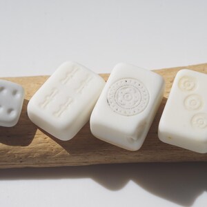 May include: A collection of white, rectangular and square-shaped beads with embossed designs. The beads are arranged on a piece of driftwood. The designs include dots, lines, and circular patterns.