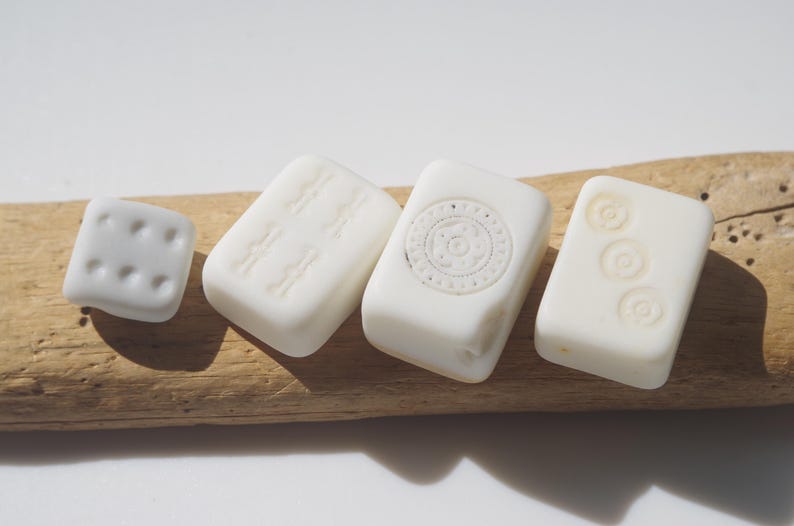 May include: A collection of white, rectangular and square-shaped beads with embossed designs. The beads are arranged on a piece of driftwood. The designs include dots, lines, and circular patterns.