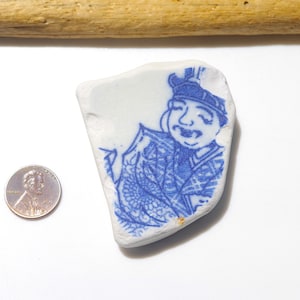 May include: A fragment of white ceramic featuring a blue illustration of a figure in traditional attire. A penny is positioned beside the ceramic. A piece of driftwood is at the top.