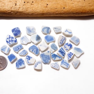 May include: A collection of small, irregular ceramic shards featuring various blue and white patterns, arranged alongside a copper-colored penny for scale. The pieces display diverse geometric, floral, and abstract designs on white backgrounds.