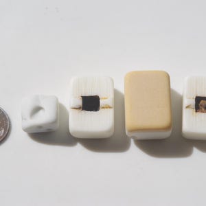 May include: A collection of handmade beads in various shapes and colors. The beads include a small white cube, rectangular white beads with dark square and brown accents, and a tan rectangular bead. A US penny is included for size reference.