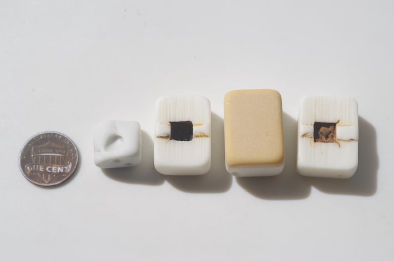 May include: A collection of handmade beads in various shapes and colors. The beads include a small white cube, rectangular white beads with dark square and brown accents, and a tan rectangular bead. A US penny is included for size reference.