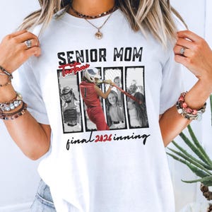 May include: White t-shirt with the text "SENIOR MOM" above a graphic of a softball player in red uniform. The graphic includes the text "Tartan" and "final 2026 inning". The shirt is a casual wear item.