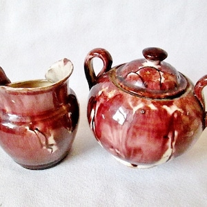 May include: A vintage ceramic creamer and sugar set in a deep burgundy color with white accents. The set has a rustic, hand-painted look.
