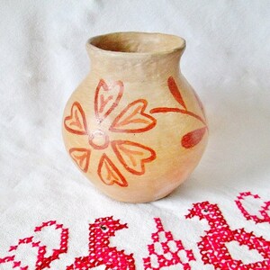 May include: A terracotta vase with a hand-painted orange flower design. The vase is a light brown color and has a rounded shape.