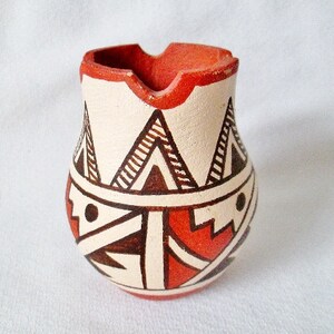 Puede incluir: Una pequeña jarra de cerámica blanca con un borde rojo y diseños geométricos en marrón y rojo. La jarra tiene un labio ligeramente acampanado y un cuerpo redondeado.