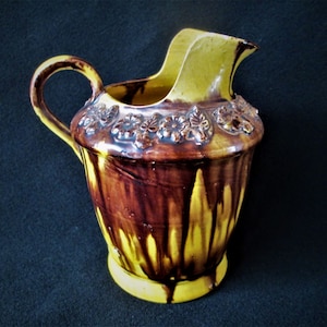 May include: A yellow and brown ceramic pitcher with a decorative floral design around the top. The pitcher has a wide mouth and a handle.