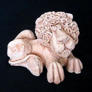 Può includere: Una scultura in ceramica di un leone con una criniera dettagliata realizzata con piccole forme arricciate. Il leone è sdraiato con la testa girata di lato e gli occhi chiusi. La scultura è dipinta in un colore beige chiaro.