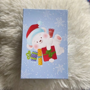 May include: A blue gift box featuring a white polar bear wearing a red Santa hat and holding presents. The box is decorated with white snowflakes.