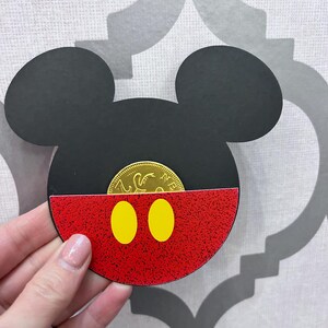 May include: A red and black Mickey Mouse head cutout with yellow circles for eyes. A gold coin is visible inside the cutout.