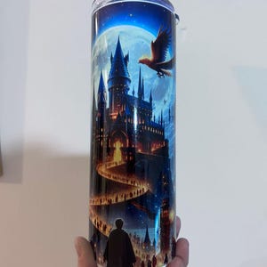 May include: A stainless steel tumbler with a detailed illustration of a castle under a full moon, with a path of people leading to the castle. A bird flies in the sky. The tumbler is held by a hand.