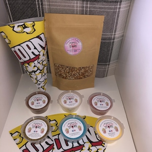 May include: A set of popcorn toppings in small containers with labels that say "Poppy's Candy Store" and a bag of popcorn kernels. The toppings include caramel, blue raspberry, strawberry, cinnamon, and yellow.
