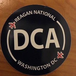 Könnte beinhalten: Ein runder Aufkleber in Blau und Weiß mit dem Text "Reagan National (DCA) Washington DC" und zwei roten Flugzeugen auf beiden Seiten des Textes.