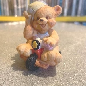 May include: A small, brown teddy bear figurine wearing a blue and white hat, riding a red tricycle with black wheels.