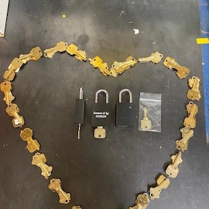 May include: A heart shape made of brass keys surrounds two black padlocks, a key, and a key-shaped tool. One padlock has the text "Suzanne & Co" and the date "25/06/23". The keys are arranged on a dark surface.