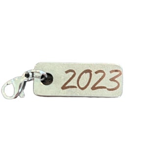 Puede incluir: Encanto de plata con un cierre de plata y una etiqueta rectangular con el número 2023 escrito en tinta marrón.