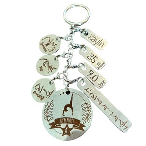 May include: Silver-colored gymnast keychain with multiple charms. The main charm features a gymnast silhouette and the word "GYMNAST". Additional charms include a name, numbers, and gymnastics poses. The keychain is attached to a silver keyring.