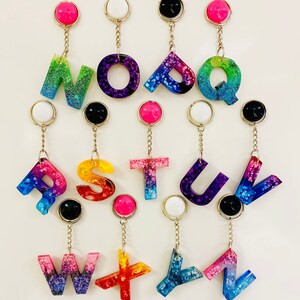Puede incluir: Un conjunto de 26 llaveros de letras de resina de colores con purpurina incrustada en la resina. Cada letra es de un color diferente y tiene una cadena dorada unida a un anillo plateado. Las letras están dispuestas en orden alfabético.