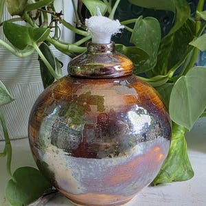 May include: A handcrafted, round oil lamp with a lid. The lamp has a glossy, glazed finish with shades of brown, bronze, and purple. A white wick extends from the top. The lamp is a decorative item for home decor.