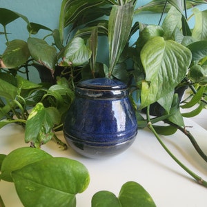 May include: A dark blue ceramic jar with a lid. The jar has a glossy finish and a subtle texture. The jar is sitting on a white surface.