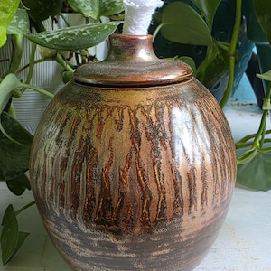 May include: A handcrafted, round oil lamp with a lid. The lamp has a textured, brown and gold finish with a white wick. The ceramic lamp is a decorative item for home decor.