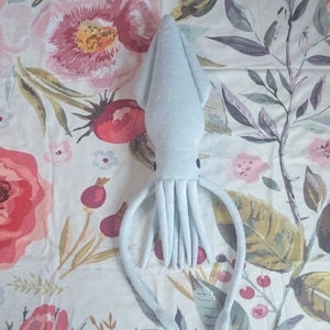 Könnte beinhalten: Ein hellblaues Plüsch-Tintenfisch-Spielzeug mit langen Tentakeln. Das Spielzeug liegt auf einem weißen Stoff mit einem Blumenmuster.