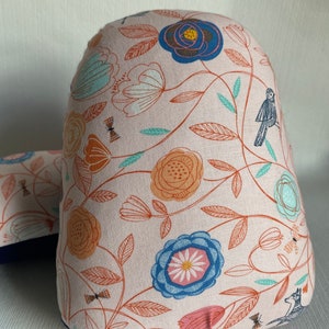 May include: A decorative cushion featuring a floral design in pink, orange, blue, and teal. The pattern includes stylized flowers, leaves, and a bird. The cushion has a rounded top and a rectangular base.