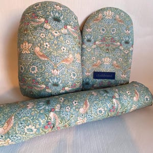 Op de afbeelding: Een set van drie teal stoffen items met een bloemen- en vogelpatroon. De set bevat twee ovenwanten en een lange, cilindrische bolster. De stof heeft een herhalend ontwerp van vogels, bloemen en bladeren.