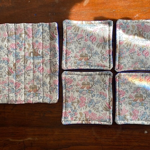 Könnte beinhalten: Ein Set aus gesteppten Stoffuntersetzern und einem passenden Tischset. Die Untersetzer und das Tischset weisen ein Blumenmuster in Rosa, Blau und Beige auf. Das Tischset ist rechteckig, und die Untersetzer sind quadratisch.