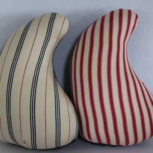 May include: Two decorative, teardrop-shaped pillows. One pillow features cream fabric with navy and burgundy stripes. The other pillow has cream fabric with red stripes. Both pillows have a dark bottom.