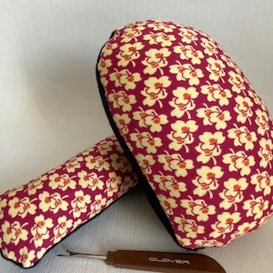 Könnte beinhalten: Ein Nähset mit einem Nadelkissen und einem kleinen Kissen, beide mit bordeauxfarbenem Stoff mit cremefarbenem Blumenmuster bezogen. Ein Nahttrenner mit braunem Griff und dem Wort "CLOVER" ist ebenfalls vorhanden. Für das Nähen.