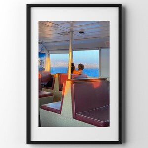 Può includere: Fotografia incorniciata di un interno di traghetto con sedili bordeaux e vista sull'oceano attraverso una finestra. Due persone sono sedute, rivolte verso l'acqua. La luce del sole crea un'atmosfera serena.