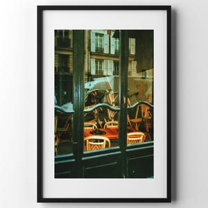 Può includere: Fotografia incorniciata di una scena di caffè parigino. L'immagine, vista attraverso un telaio di finestra verde scuro, mostra sedie e tavoli di legno, con edifici sullo sfondo. La scena è immersa in una luce calda e naturale.