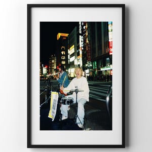 Può includere: Fotografia incorniciata di un musicista di strada che suona la batteria in una via della città di notte. Il musicista indossa una felpa bianca e pantaloni chiari. Lo sfondo presenta edifici e insegne illuminate.
