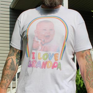 May include: White t-shirt featuring a baby graphic within a rainbow arch. The text "I LOVE GRANDPA" is printed in colorful letters below the image. The shirt is a casual, everyday wear item.