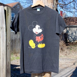 May include: Black t-shirt with a vintage Mickey Mouse graphic. Mickey is wearing red shorts and yellow shoes.