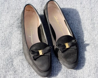 May include: A pair of black suede loafers with a gold buckle and a black bow. The loafers have the text "Salvatore Ferragamo" printed on the inside.