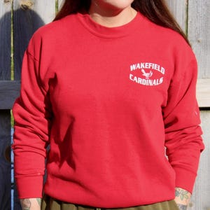 May include: A red crew neck sweatshirt with the words "Wakefield Cardinals" in white arched text above a bird graphic. The long-sleeved top is made of a soft material.