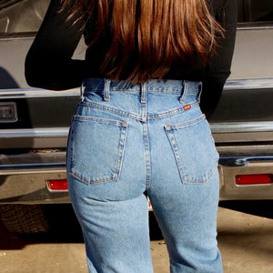 May include: A person wearing a black long-sleeved shirt and blue denim jeans with a high waist. The person is standing in front of a car.