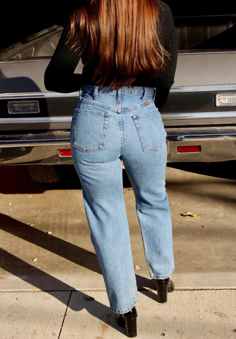 May include: A person wearing a black long-sleeved shirt and blue denim jeans with a high waist. The person is standing in front of a car.