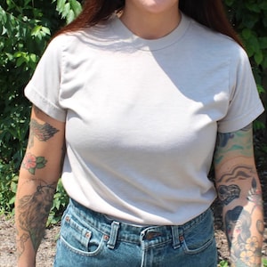 May include: A light gray, short-sleeved t-shirt. The shirt is made of a soft material and is paired with blue denim jeans. The person wearing the shirt has multiple tattoos on their arms. The background features green foliage.