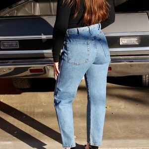 May include: A person wearing a black long-sleeved shirt and blue denim jeans with a high waist. The person is standing in front of a silver car.