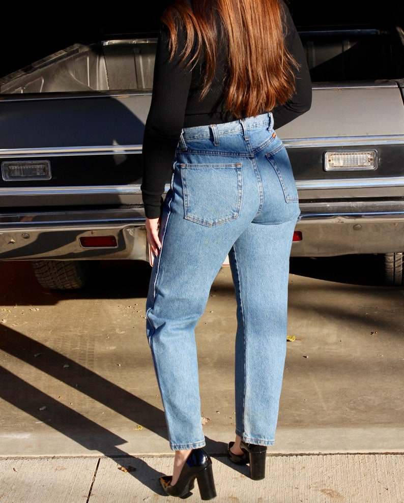 May include: A person wearing a black long-sleeved shirt and blue denim jeans with a high waist. The person is standing in front of a silver car.