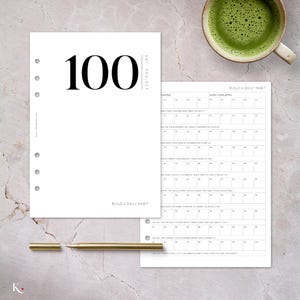 Könnte beinhalten: Ein weißer Planer mit der Zahl "100" in großer schwarzer Schrift und dem Text "DAY PROJECT". Eine Seite "Build a Daily Habit" ist sichtbar. Ein goldener Stift liegt in der Nähe des Planers, und eine Tasse grüner Tee ist im Hintergrund.