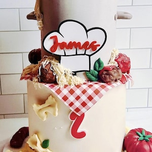 May include: A two-tiered cake decorated with a chef's hat, the name "James", and various Italian-themed decorations, including spaghetti, meatballs, and a red checkered tablecloth. The bottom tier has a large number "1" on it.
